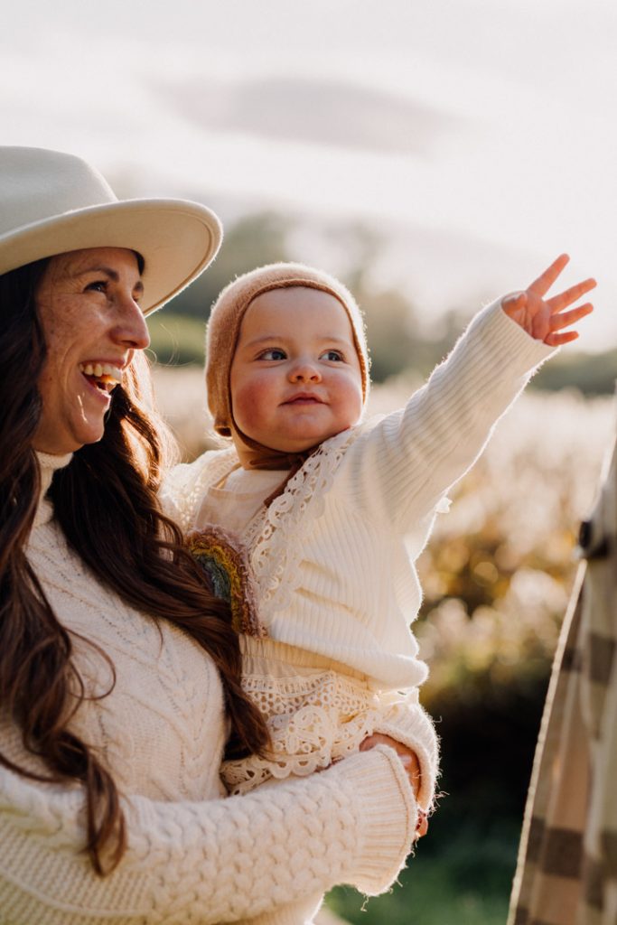Little one year old girl is reaching out her hand and smiling. Mum is holding her and smiling too. Hampshire photo session celebrating one year old. Golden hour lifestyle photo shoot. Ewa Jones Photography
