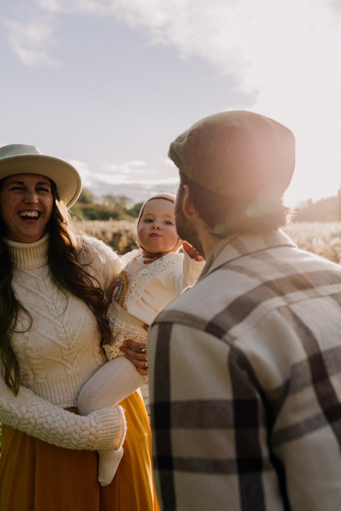Parents share a joyful laugh while holding their baby during an outdoor family photo session, with soft sunlight and tall grasses creating a warm, golden-hour backdrop. Winchester photo shoot. Hampshire photographer. Ewa Jones Photography