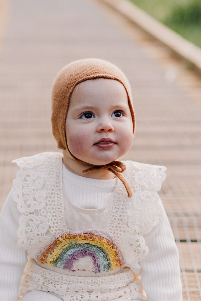 one year old girl is looking up. she is sitting on the wooded path. girl is wearing lovely outfit with rainbow on her top. Hampshire photo shoot. Celebrating one year old with an outdoor photo session. Hampshire photo shoot. Ewa Jones Photography