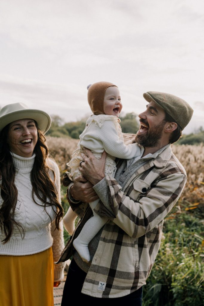 Father lifts and laughs with his baby during an outdoor family photo session, while the mother smiles nearby in a natural field setting under soft, overcast light. Golden hour photo session. Hampshire photo shoot. Ewa Jones Photography