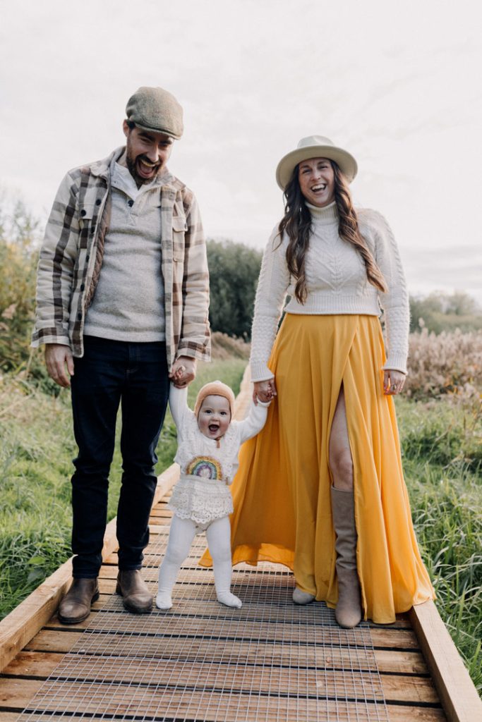 Parents hold their baby’s hands as the child takes steps along a wooden path during an outdoor family photo session, all smiling and laughing in a natural field setting. Celebration of one year old. Natural outdoor family photo session. Hampshire photographer. Ewa Jones Photographer