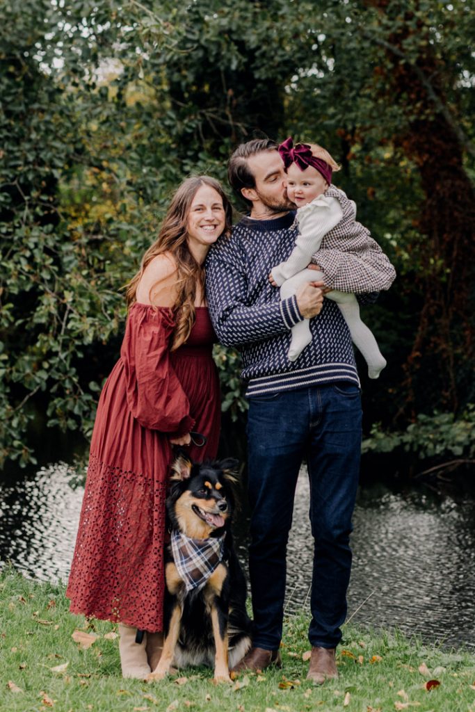 Family of four is standing next to the river and enjoying cuddles. Mum is wearing nice dress and their girl is looking away. lifestyle and natural photo session in Hampshire. Ewa Jones Photographer