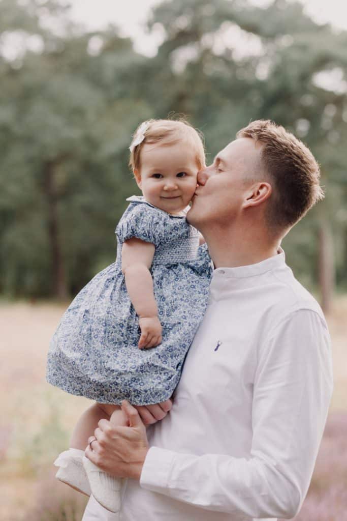 Dad is holding his daughter and is giving her a kiss in her cheek. Natural outdoor family photo shoot in Berkshire. Family photographer in Hampshire. Ewa Jones photography