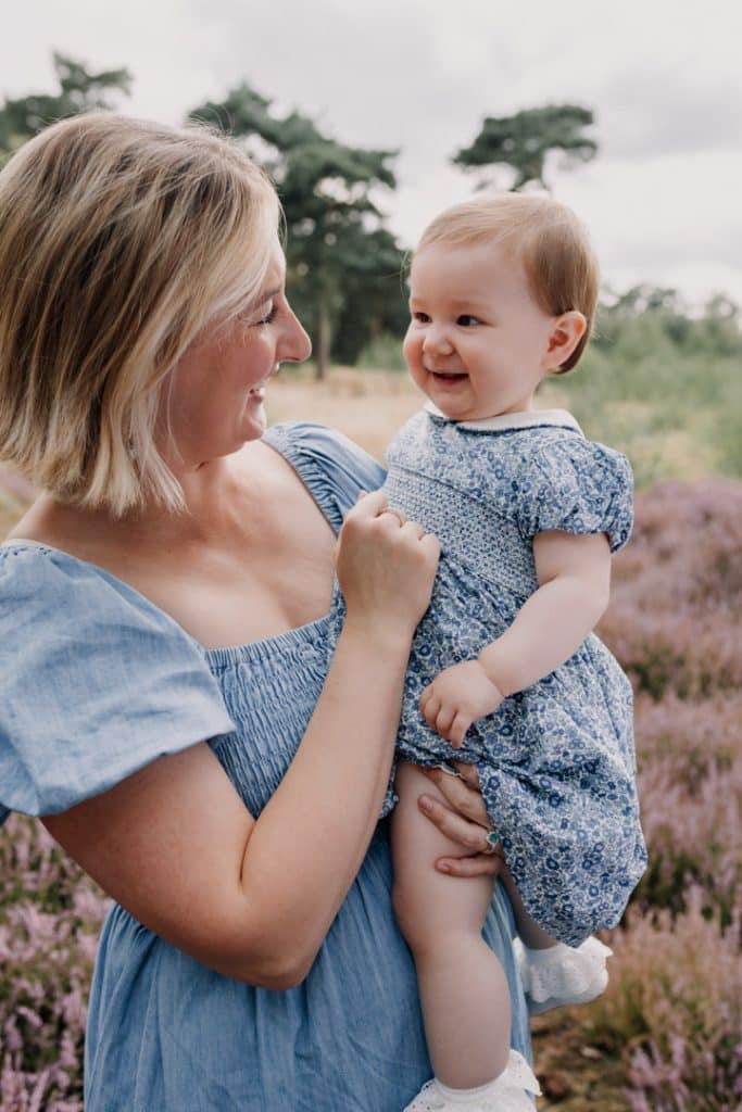 Mum is holding her daughter and looking at her. The girl is looking back at mum and smiling. Family photo session in Berkshire. Mum is wearing lovely blue dress and her daughter has a blue dress too. Hampshire photographer. Ewa Jones Photography