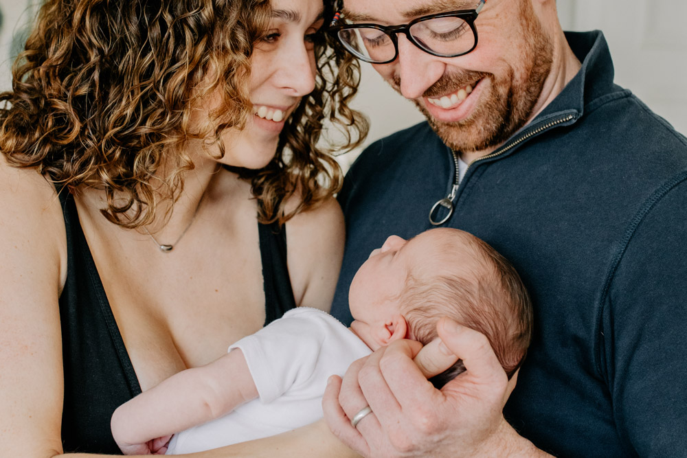 Close up of family cuddling with each other. mum is holding baby and looking at her husband. husband is smiling and looking down at his baby. Hampshire in home newborn photography. Ewa Jones Photography