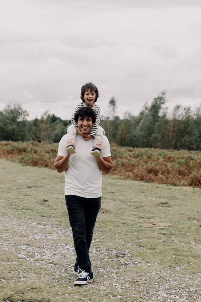 Dad is holding his son on his shoulders and both are smiling and walking towards the camera. Hampshire family photo shoot. Family photographer. Ewa Jones Photography