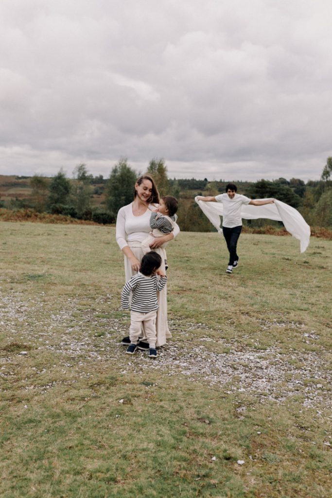 Mum is holding her son and other son is looking at his dad. dad is running towards them trying to catch them. family poses during family photo session. Ewa Jones Photography
