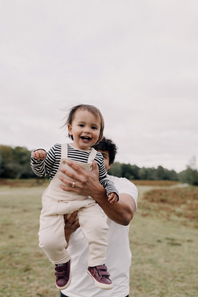 Dad is lifting his son towards the camera. little boy is smiling. he is wearing white dungarees and stripy top. family fun during family photo session. natural photographs. Ewa Jones Photography