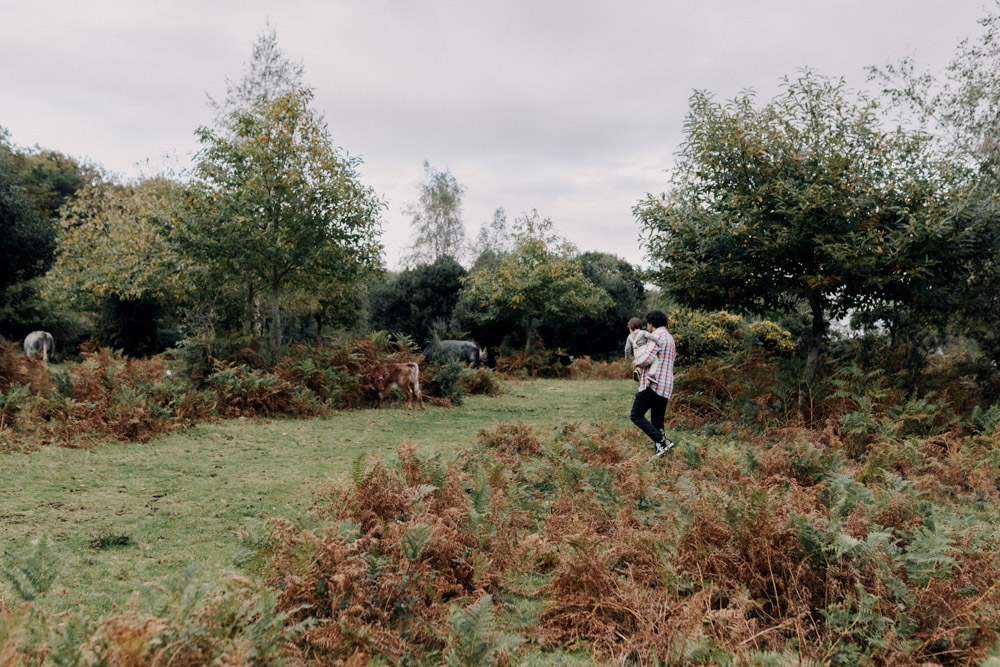 dad is holding his son and walking towards wild horses in the New Forest. Hampshire family photo shoot. Family photographer. Ewa Jones Photography
