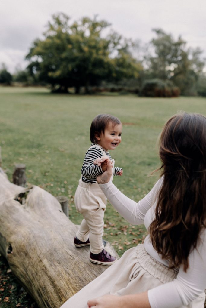 little boy is standing on the log and smiling. Mum is holding his hand. Candid family photo session. Ewa Jones Photography