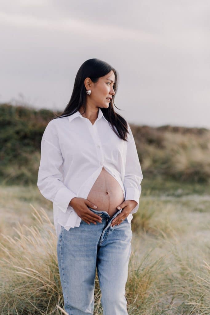 Expecting mum is wearing jeans and white shirt and looking away. she is standing at the beach. Maternity photo shoot at the beach. Hampshire maternity photographer. Ewa Jones Photography