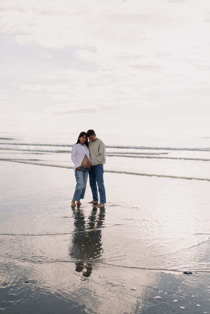 couple is standing on the sand and cuddling. mum is wearing white shirt and jeans, dad to be is wearing jeans and a jumper. Hampshire pregnancy photo session. Ewa Jones Photography