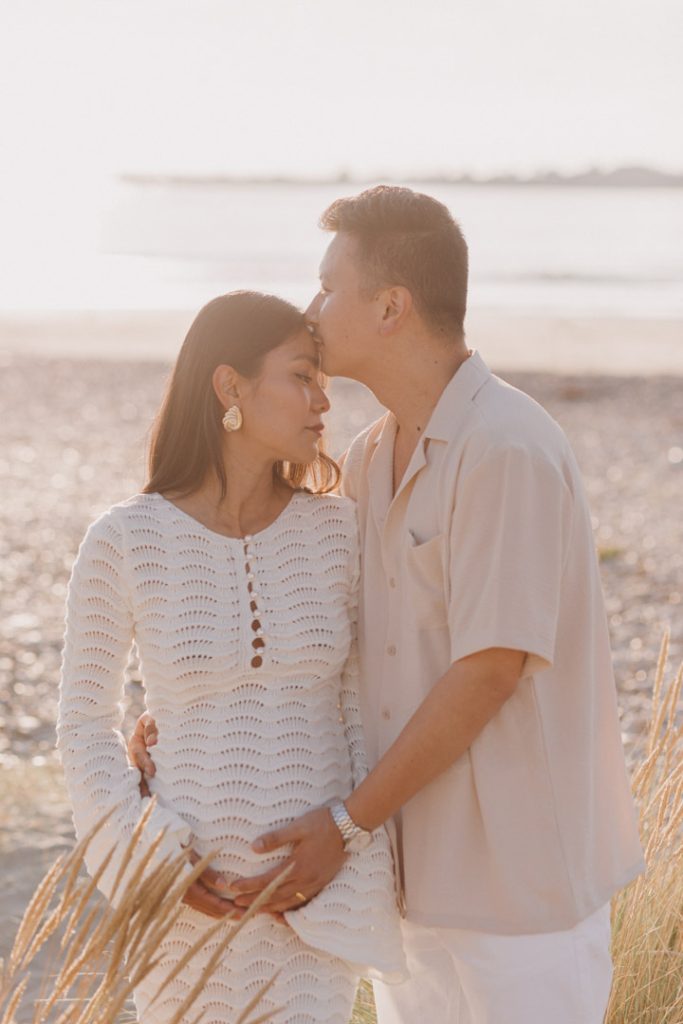 dad is cuddling close to his wife and kissing her in her temple. Maternity photo shoot at the beach. mum is wearing a white dress and dad is wearing shirt and white trousers. Ewa Jones Photography