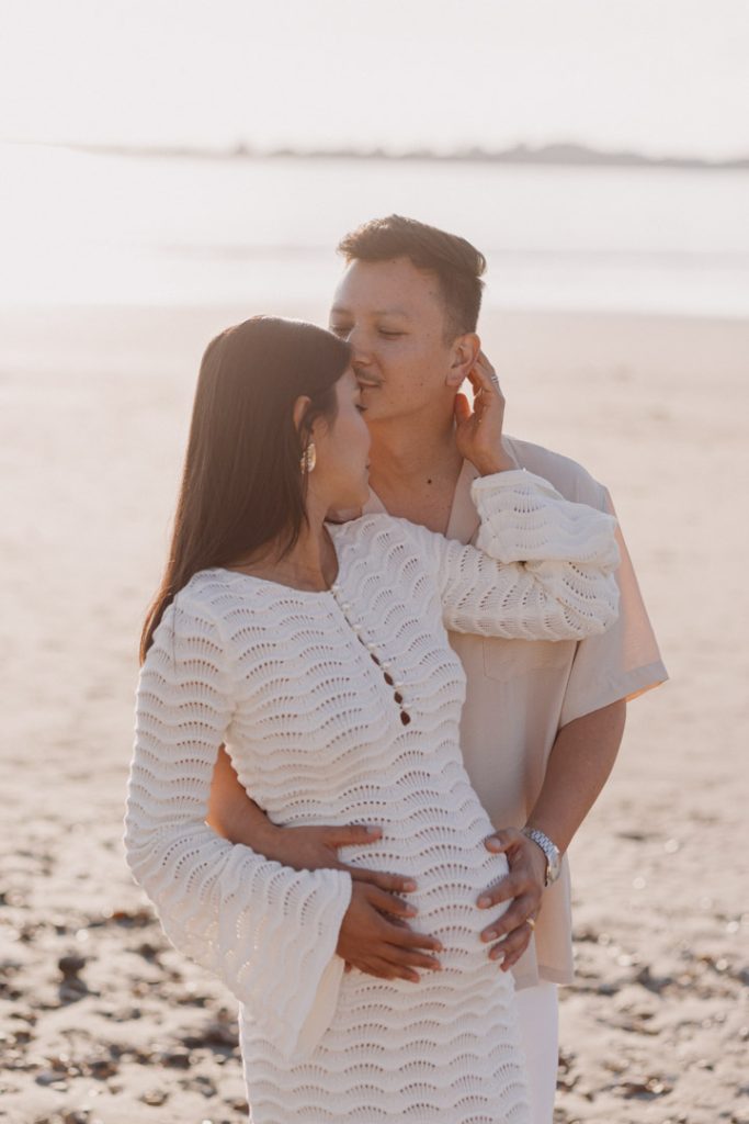 dad is cuddling close to his wife and kissing her in her temple. Maternity photo shoot at the beach. mum is wearing a white dress and dad is wearing shirt and white trousers. Ewa Jones Photography