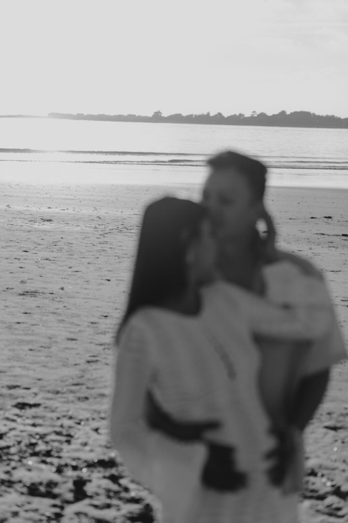 dad is cuddling close to his wife and kissing her in her temple. Maternity photo shoot at the beach. mum is wearing a white dress and dad is wearing shirt and white trousers. Ewa Jones Photography