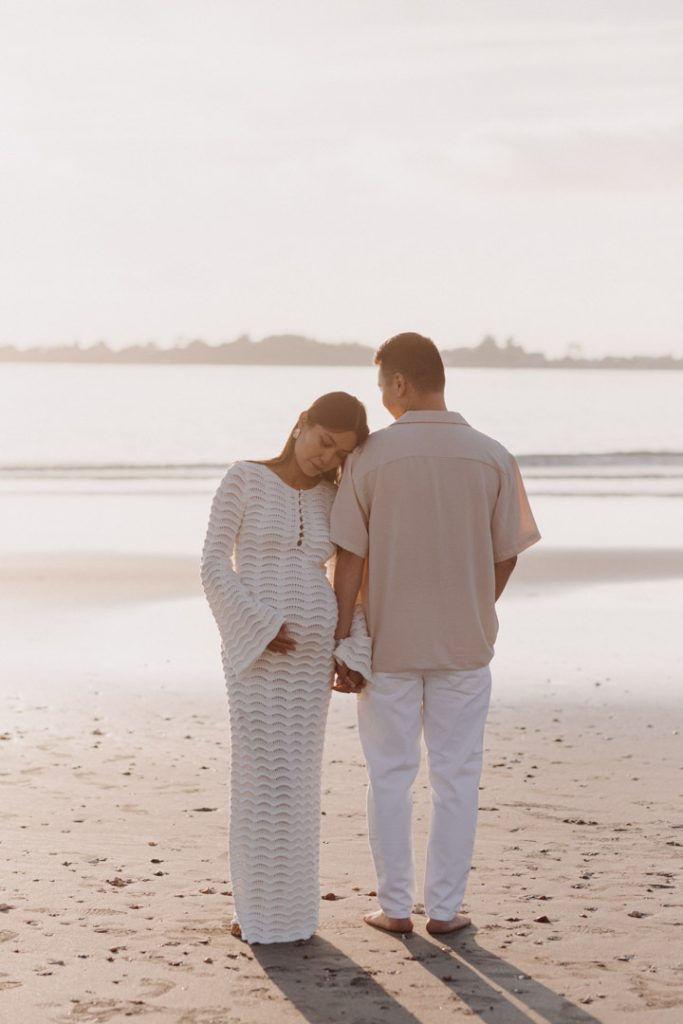 Mum to be is looking down at baby bump and holding the bump. her husband is standing away from the camera and looking at the sea. Maternity photo session at the beach. Hampshire bump photographer. Ewa Jones Photography
