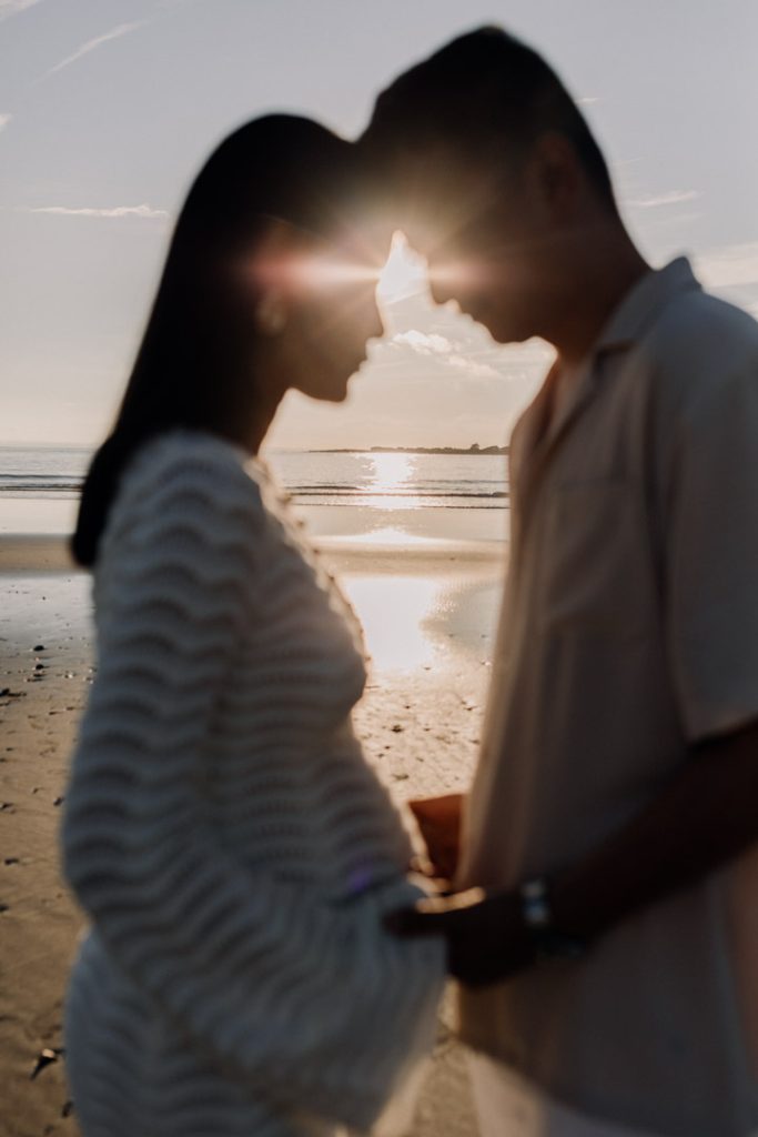 A softly blurred silhouette of two people standing face-to-face on a beach at sunset, with their foreheads touching. The sun shines directly between their faces, creating a warm starburst effect. They are holding hands over a visibly pregnant belly, suggesting an expectant couple. Gentle waves, wet sand reflecting the sunlight, and a calm horizon form the background, giving the image a peaceful, intimate, and romantic atmosphere. Ewa Jones photography