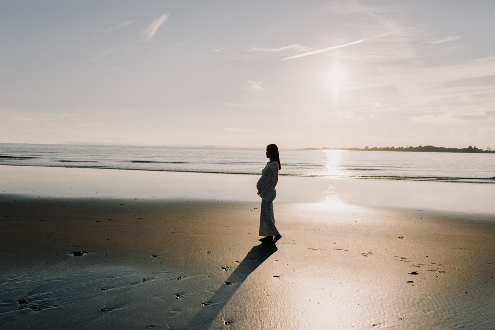 A pregnant person stands in profile on a quiet beach at sunset, gently cradling their baby bump while facing the ocean. Wearing a long white textured dress, they are silhouetted against the warm light reflecting across the calm water and wet sand. A long shadow stretches behind them, with a distant shoreline and soft, hazy sky creating a serene, contemplative atmosphere.. Maternity photographer in Hampshire. Ewa Jones Photography