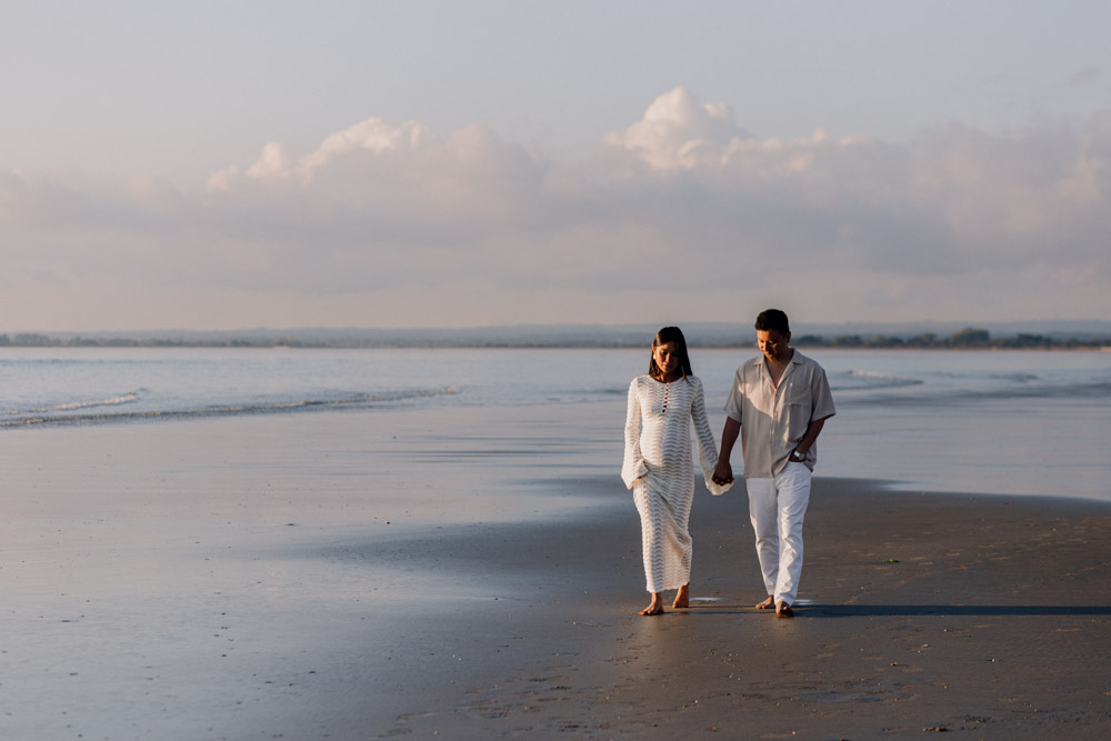 An expectant couple walks hand in hand along a quiet beach during golden hour. The pregnant person wears a long white textured dress, while their partner is dressed in a light short-sleeved shirt and white pants. Both look downward as they stroll barefoot on the wet sand near the shoreline. Calm water, soft reflections, and large pastel clouds stretch across the horizon, creating a peaceful, intimate atmosphere. Hampshire maternity photo shoot. Ewa Jones Photography. Must-Have Poses for a Natural Maternity Photo Session