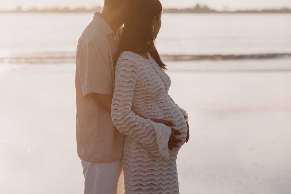An expectant couple stands together on a beach at sunset, with one partner embracing the other from behind. The pregnant person, wearing a long white textured dress, gently cradles their baby bump while their partner wraps an arm around them and rests a hand over their belly. Both face the ocean, illuminated by soft golden light reflected across the calm water, creating a warm, peaceful, and intimate moment. Hampshire newborn photographer. Ewa Jones Photography