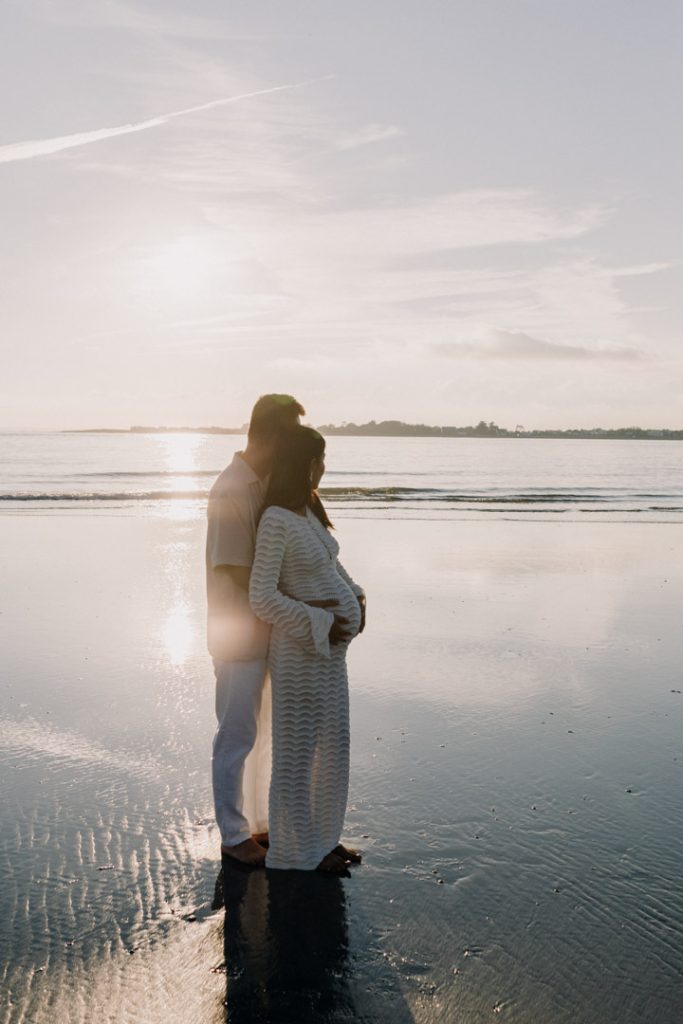 couple is standing on the water and looking into the sea. mum is holding baby bump. lovely maternity photo session at the beach. bump photo shoot. Ewa Jones Photography