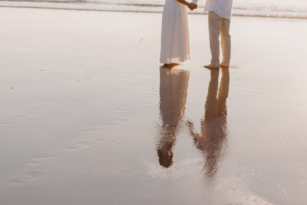 The lower half of an expectant couple stands barefoot on wet sand at the edge of a beach, holding hands during golden hour. One person wears a flowing white dress, while the other wears light-colored pants. Their elongated reflections stretch across the smooth, reflective sand, with gentle waves in the background creating a calm, romantic atmosphere. Hampshire and Basingtoke photographer. Ewa Jones Photography