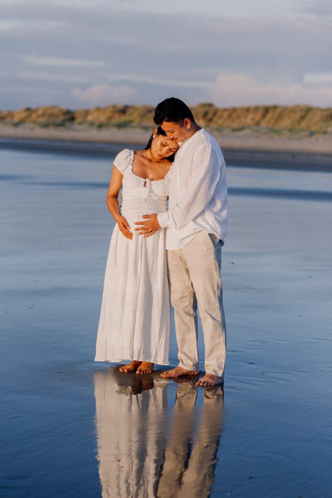 Expecting parents are holding close and looking down at the bump. lovely sun is shining on their bodies. lovely maternity photo session during golden hour at the beach. Hampshire maternity photographer. Ewa Jones Photography