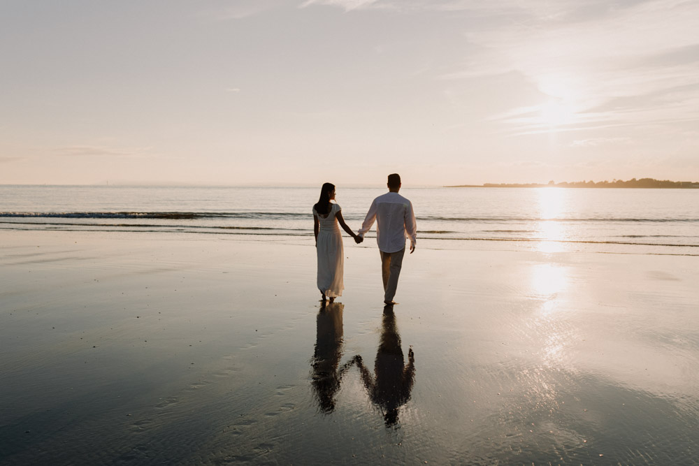 An expectant couple walks hand in hand along a quiet beach during golden hour. The pregnant person wears a long white textured dress, while their partner is dressed in a light short-sleeved shirt and white pants. Both look to the sea as they stroll barefoot on the wet sand near the shoreline. Calm water, soft reflections, and large pastel clouds stretch across the horizon, creating a peaceful, intimate atmosphere. Hampshire maternity photo shoot. Ewa Jones Photography