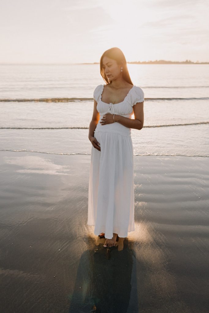 Mum is standing on the water and holding bump. she is wearing white dress. sun is shining behind her. Hampshire bump photo shoot. Ewa Jones Photography