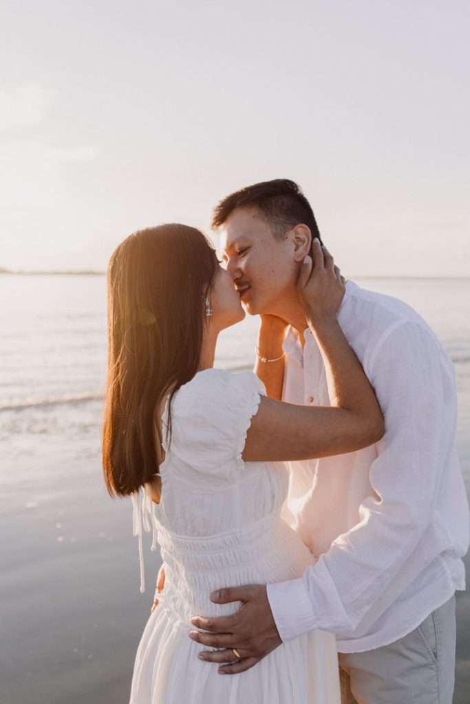 Parents to be are kissing at the beach. both wearing white outfits. What to wear for a maternity photo session. Ewa jones Photography