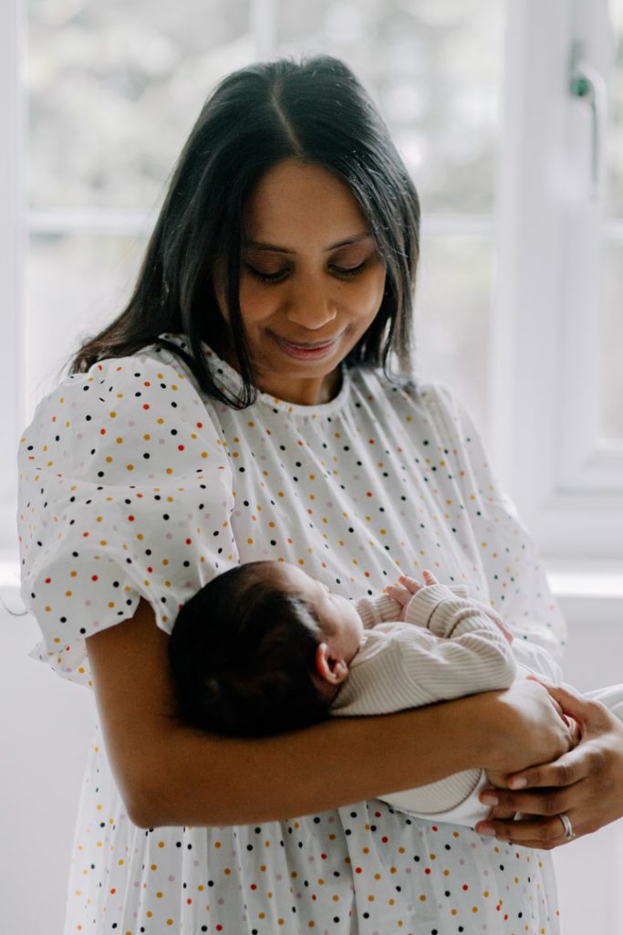 Mum is standing by the window and holding her newborn baby in her arms. in home natural newborn photo shoot. Newborn Hampshire photographer. Ewa Jones Photography