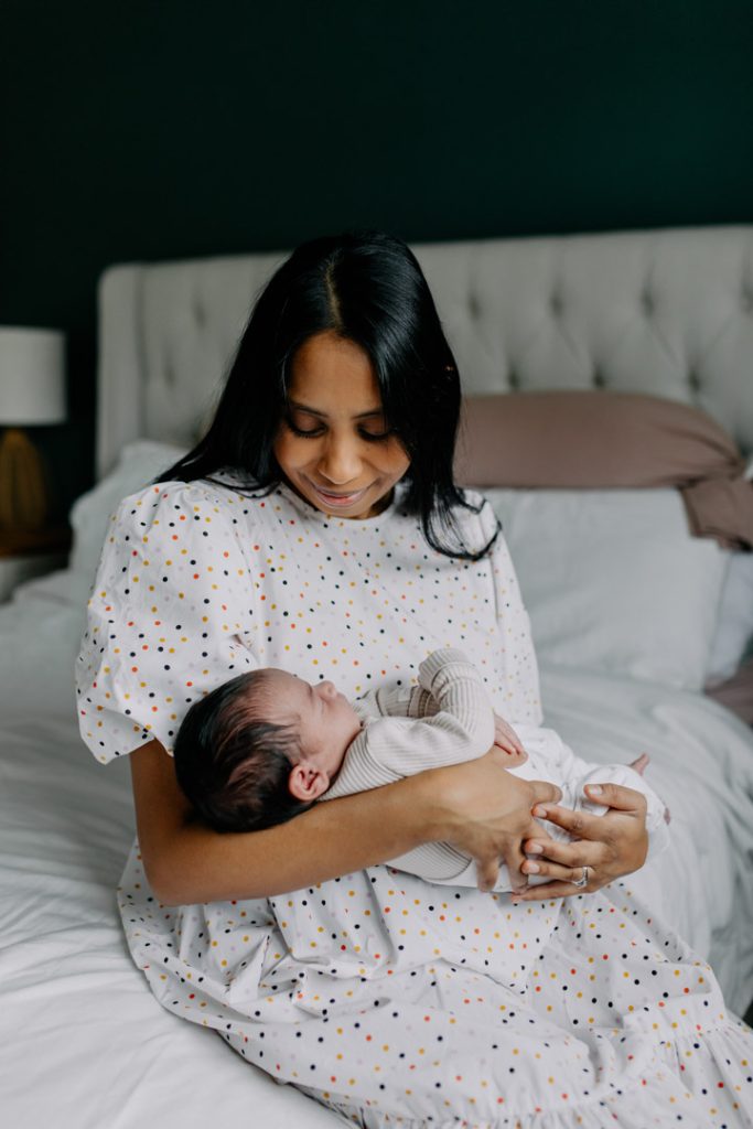 Mum is sitting at the edge of bed and looking down at her newborn baby. Basingstoke newborn photographer. Ewa Jones Photography