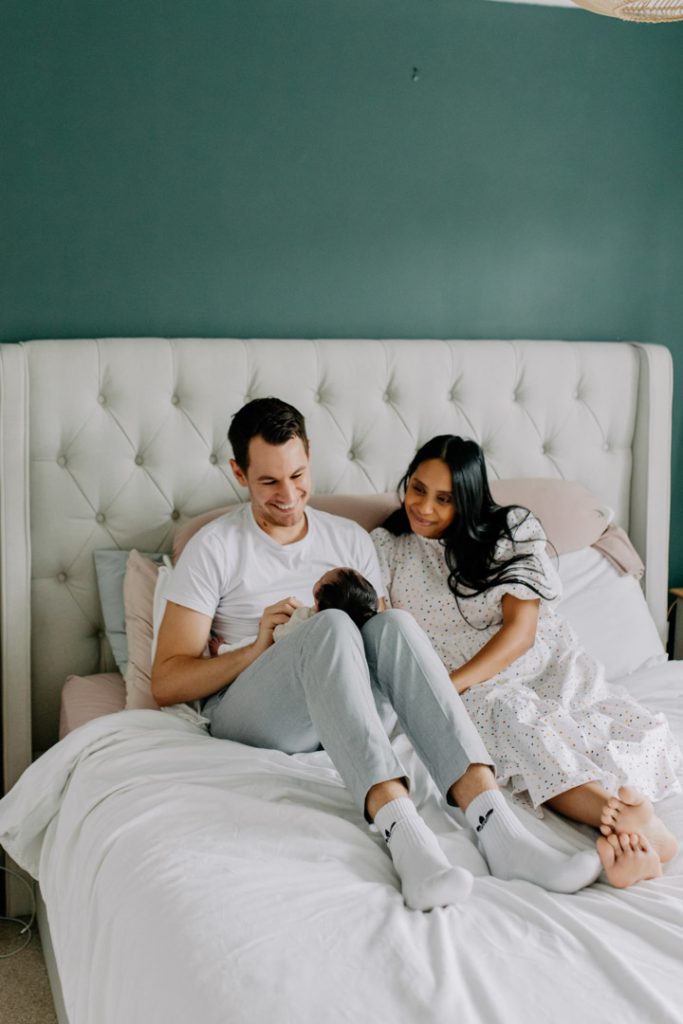 Dad and mum are sitting on bed and looking and smiling at their baby. in home newborn photo session. Ewa Jones Photography