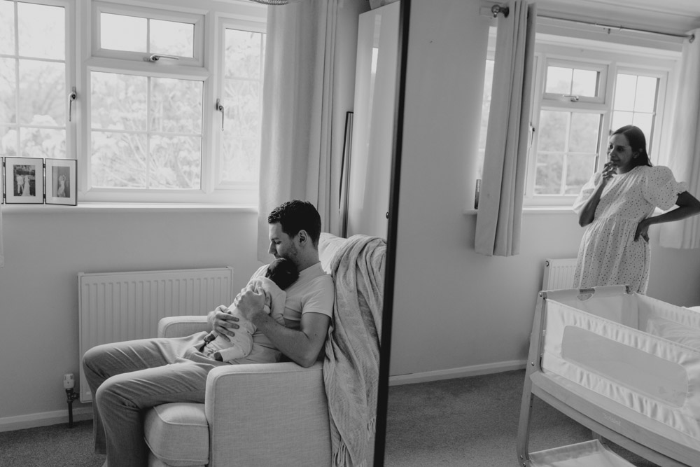 Dad is holding baby and sitting on the chair. mum reflection is in the mirror. lovely capturing the moment photo session. Newborn photo shoot in Hampshire. Ewa Jones Photography